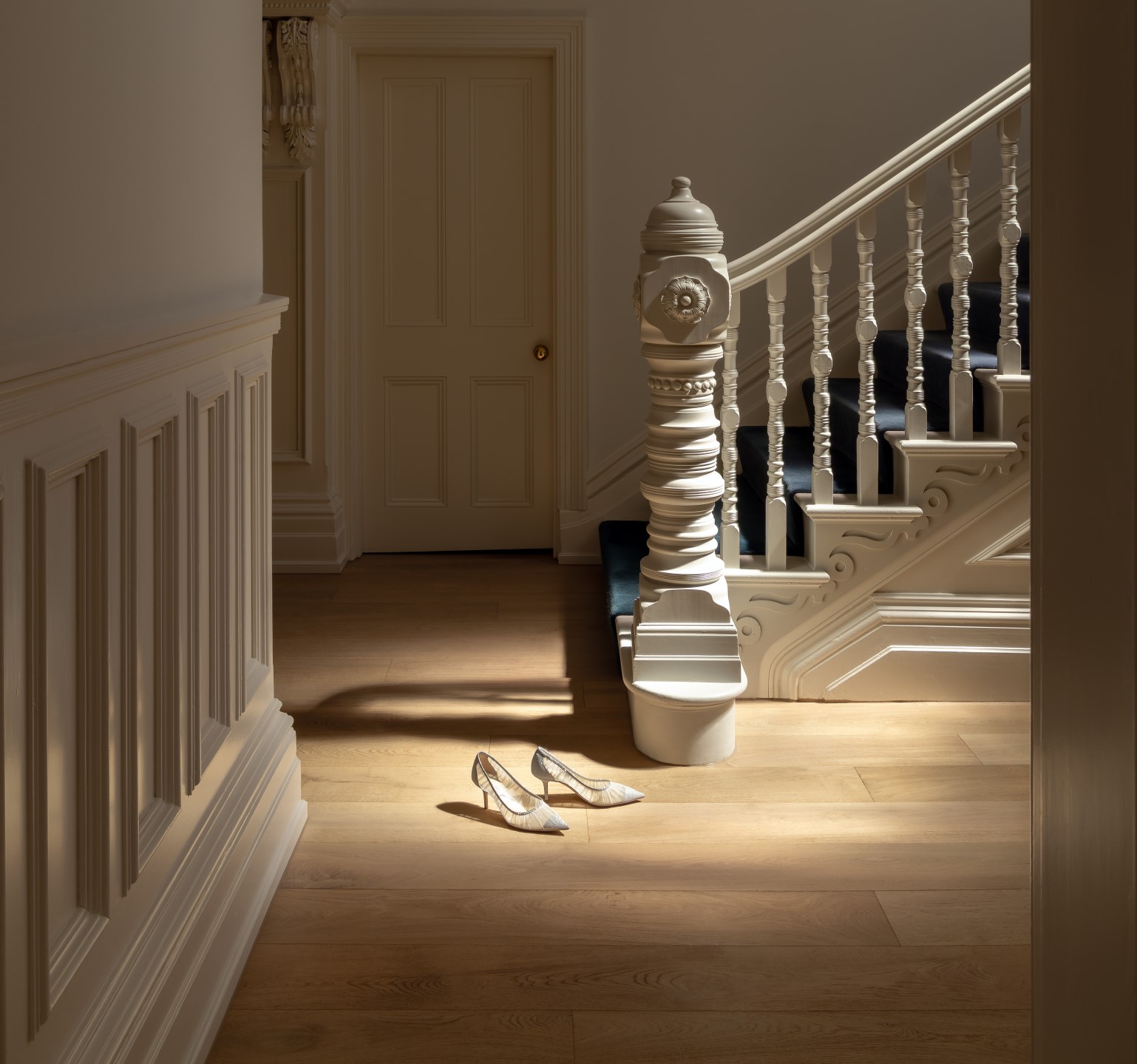 Elegant interior featuring sandy oak engineered wood flooring, illuminated by soft natural light. The space includes a grand staircase with intricate white balusters and a modern chandelier hanging from the ceiling. A pair of delicate high heels are placed on the floor, adding a touch of sophistication to the scene