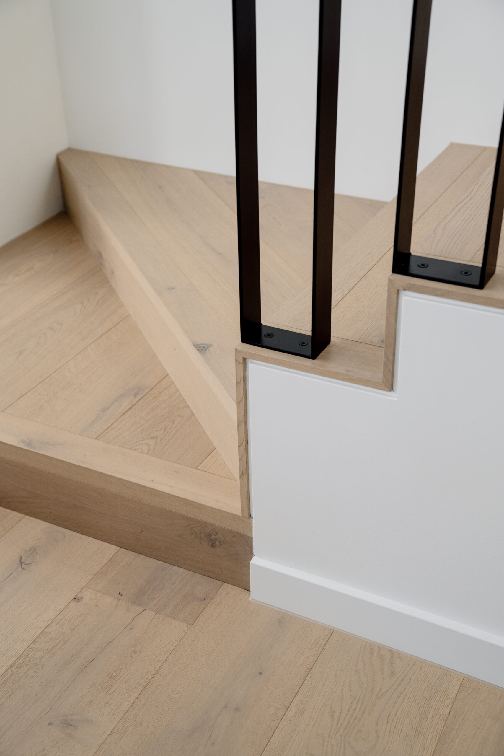 Close-up view of a staircase corner with natural oak engineered wood flooring, featuring clean lines and black metal balustrades. The image highlights the seamless integration of the flooring with the stair design, showcasing the natural wood grain and smooth finish