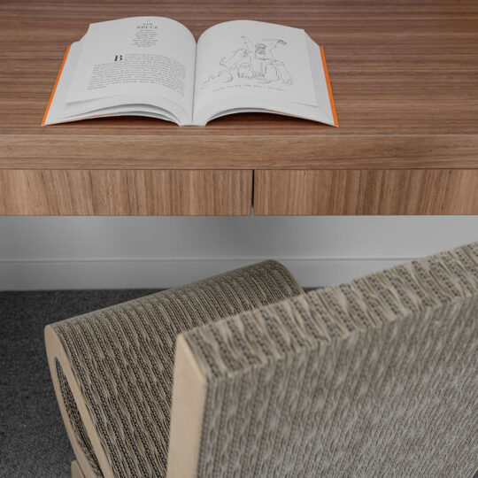 A close-up of a study area with a desktop crafted from Lignapal Blackbutt veneer. An open book rests on the desk, while a textured chair sits in front of it. The warm tones of the Blackbutt veneer provide a cozy contrast to the neutral-colored chair and gray carpet.