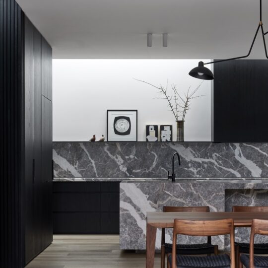 A minimalist kitchen featuring warm grey timber flooring with oversized oak floorboards, dark cabinetry, and a striking marble island. The space is defined by clean lines and a muted color palette.