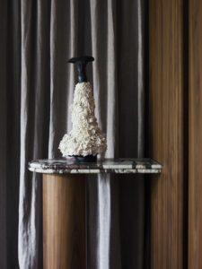 A sculptural side table with a marble top and cylindrical walnut base accompanies Lignapal walnut veneer paneling at Finch House. A beautiful sculpture sits on top of the table against an elegant drapery backdrop.